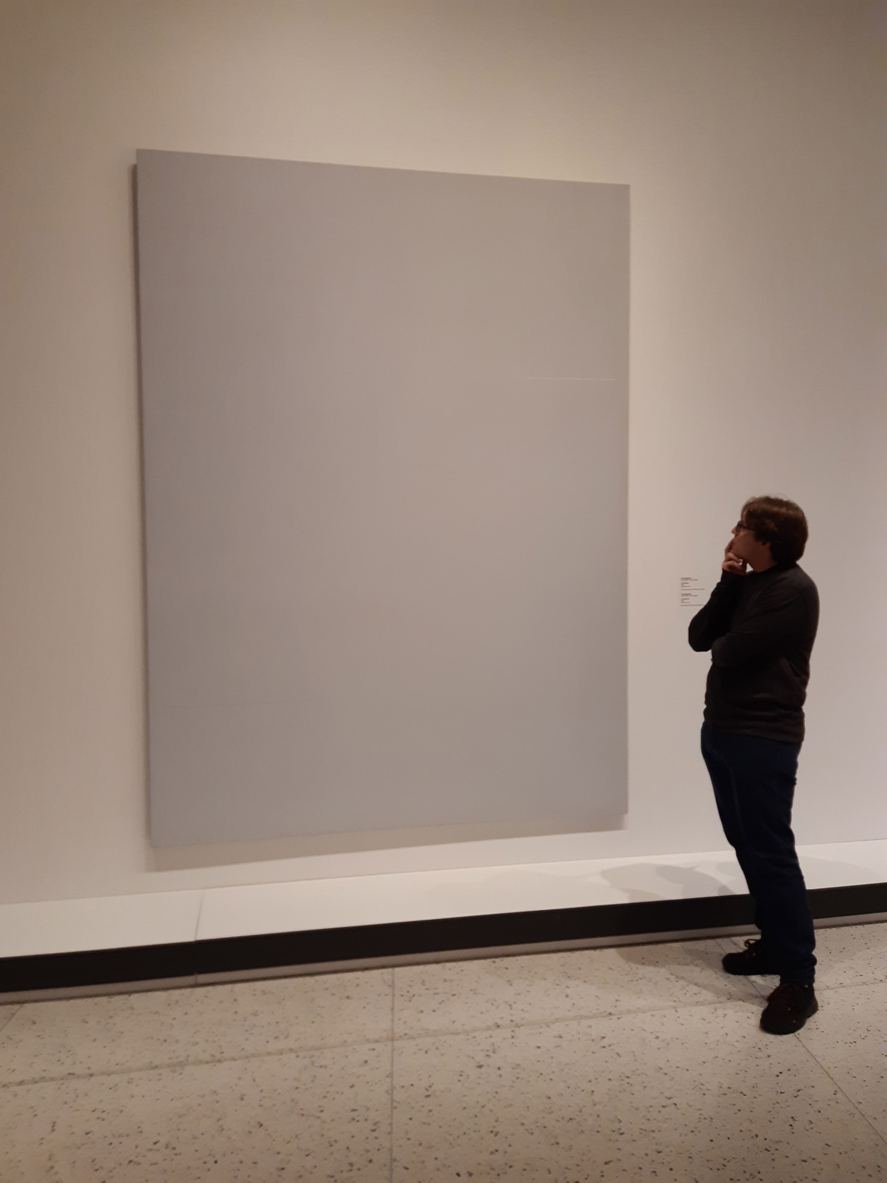 Eu, em pé, com pose pensativa, em frente a um quadro em branco